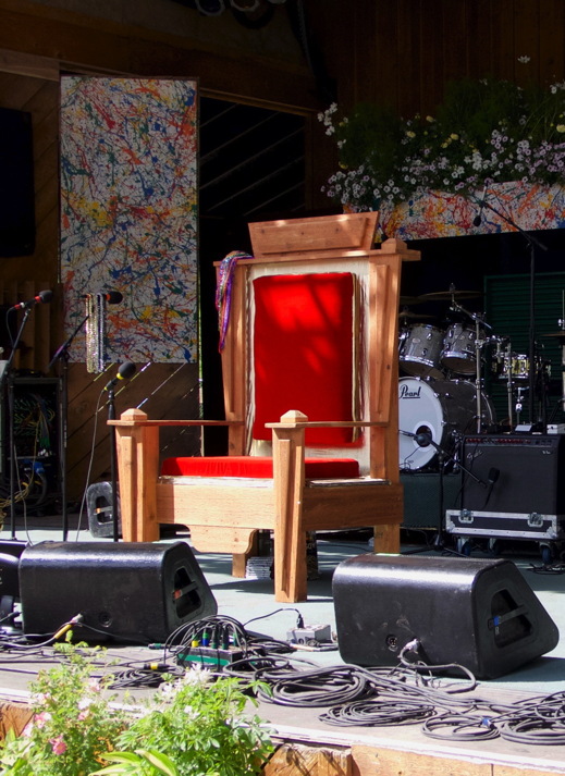A cedar throne with red fabric on a stage, with a drumset behind it and speakers and cables in front of it.